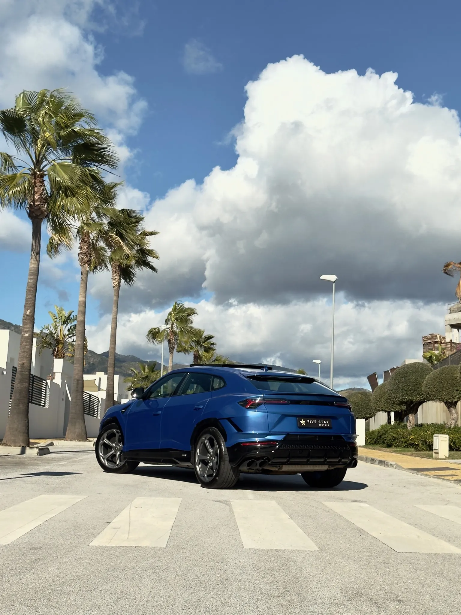 Lamborghini Urus S in Blu Eleos, rear three-quarter view with Five Star Rentals plate, Marbella palm trees and sunny sky