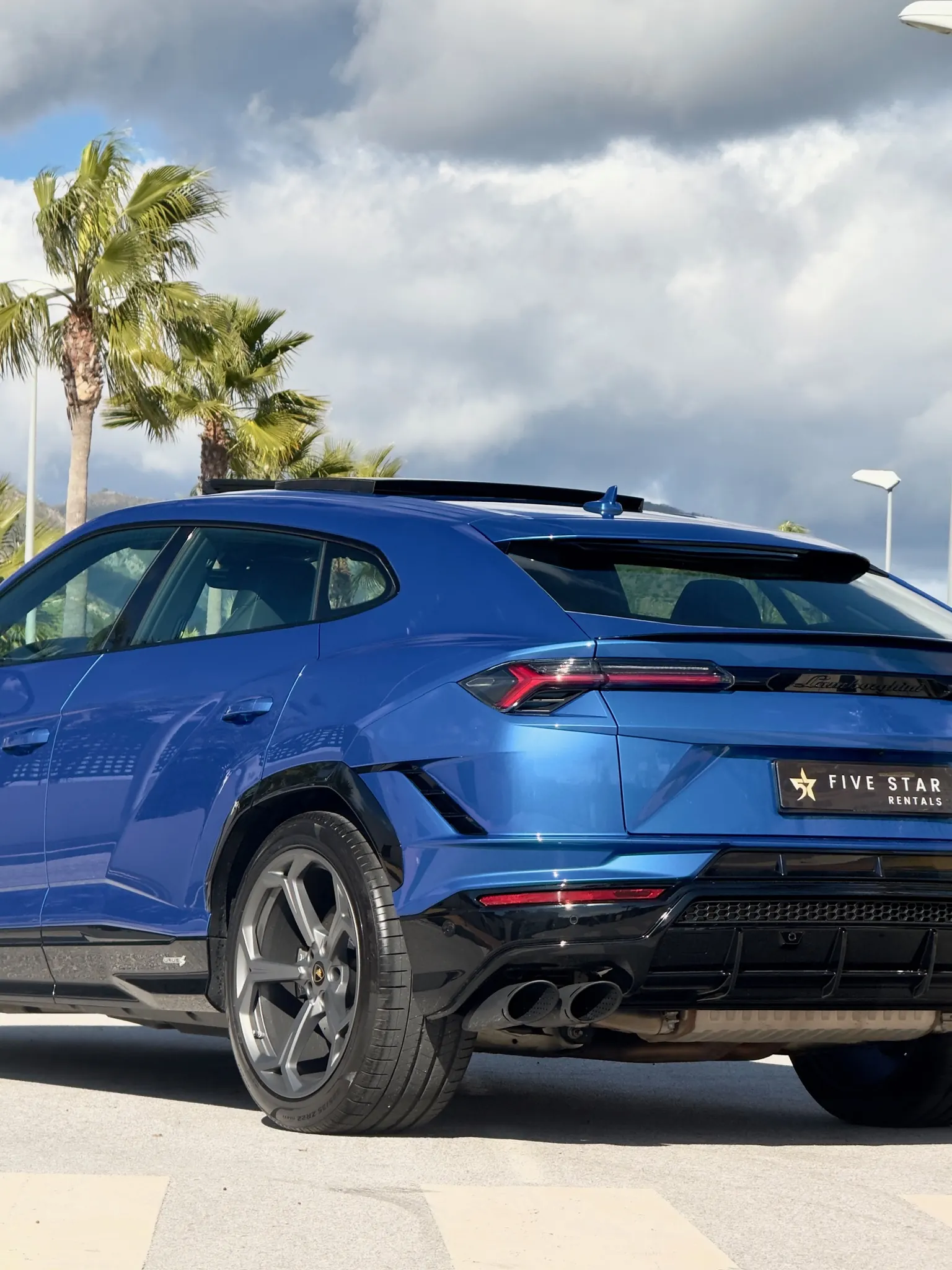 Rear side profile of the Blu Eleos Lamborghini Urus S with 22-inch titanium grey wheel, cursive Lamborghini badge and dual exhaust