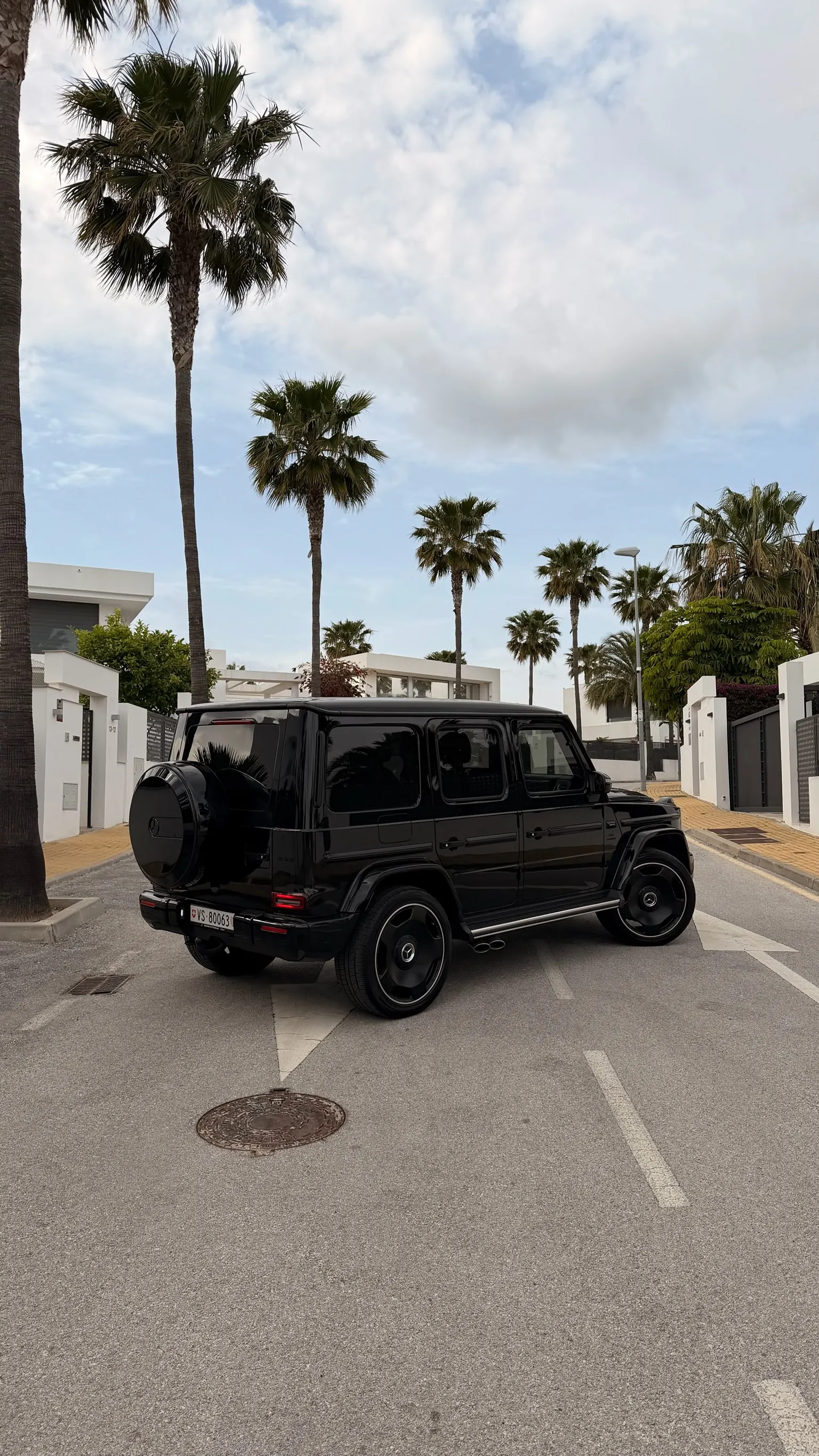 Rear three-quarter view of the Obsidian Black Mercedes-AMG G63 with body-coloured spare wheel cover and Marbella palm trees in background