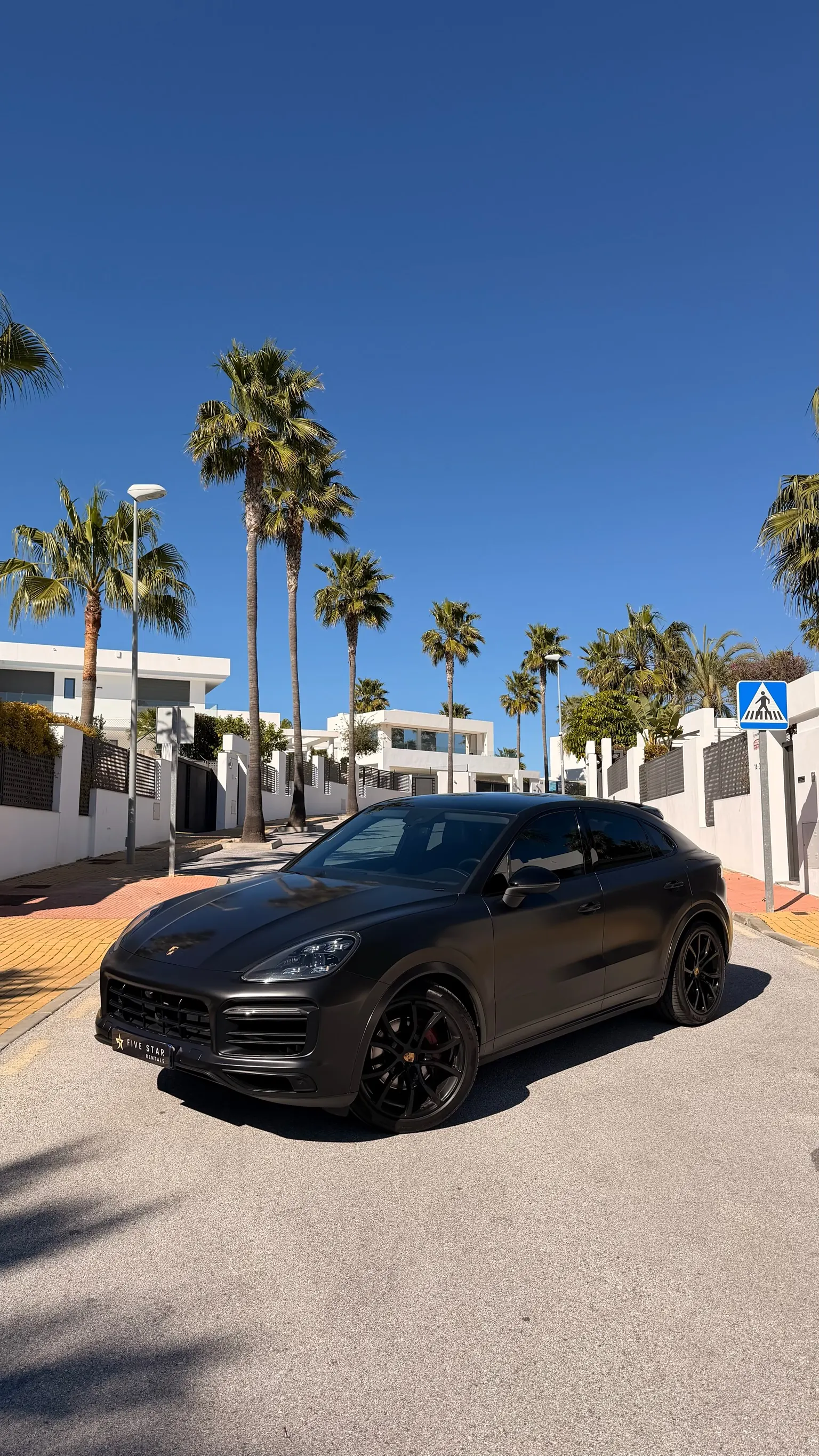 Matte black Porsche Cayenne Coupé E-Hybrid, front three-quarter view with Five Star Rentals plate and Marbella palm trees